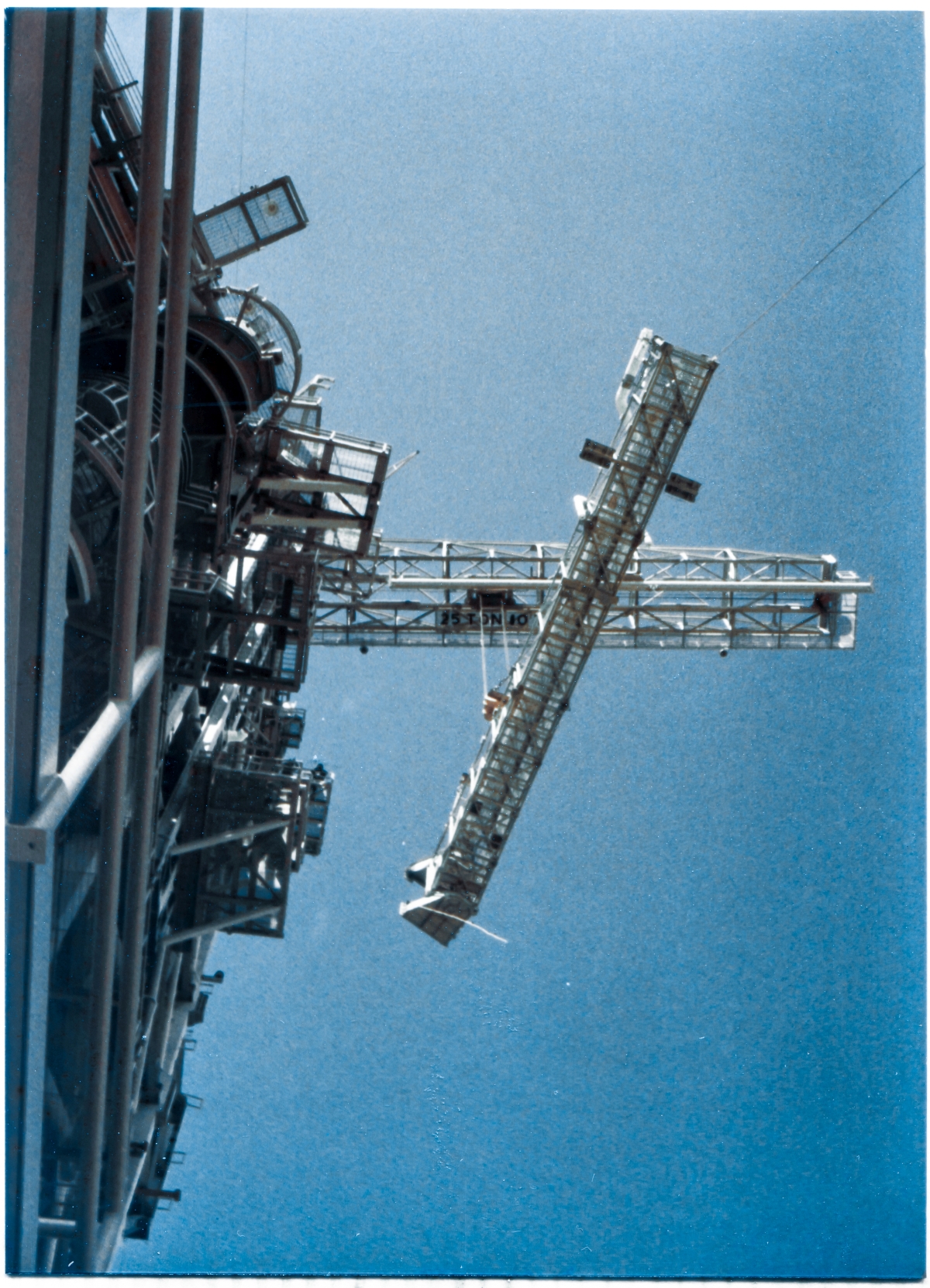 Image 108. I'm not even sure if I can sensibly describe this image, which speaks very well for itself, which is fortunate. At Space Shuttle Launch Complex 39-B, Kennedy Space Center, Florida, the GOX Arm hangs against a clear blue sky, directly overhead, suspended from the Hammerhead Crane which you also see projected against that clear blue sky above it. You are standing in a DANGEROUS place. You are standing beneath a suspended load. And a Great Slow Dance of Steel is being performed in the air above you, involving a tonnages that would wreak untold havoc should any of it get loose, fall, or otherwise behave in any way not exactly according to plan. Ivey Steel Erector's Union Ironworkers are in complete control of the situation, and all is well, but the energy compressed into what you're seeing here is beyond imagining, and it must never be allowed to escape uncontrolled. We are standing just east of MLP Mount Mechanism Number 6, on the west side of the Flame Trench, and although we are not exactly, precisely, beneath the GOX Arm, we are very very close to that point. Too close. The Lift has proceeded a little beyond the half-way point, and the Arm is roughly 150 feet above us, but it still has another hundred vertical feet to go before it reaches its destination at the top of the FSS, where it will be bolted and welded to the tower. Photo by James MacLaren.